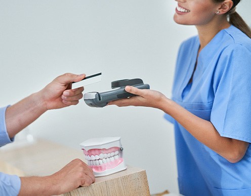 Woman in scrubs holding card reader for man’s payment at desk with artificial teeth